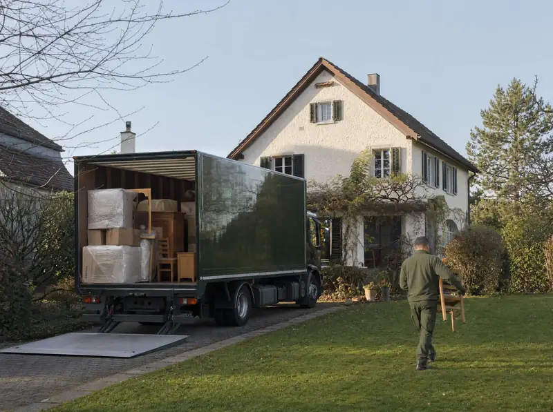 Dunkelgrüner Lastwagen vor einem Einfamilienhaus, ein Mitarbeiter trägt einen Stuhl über den Rasen